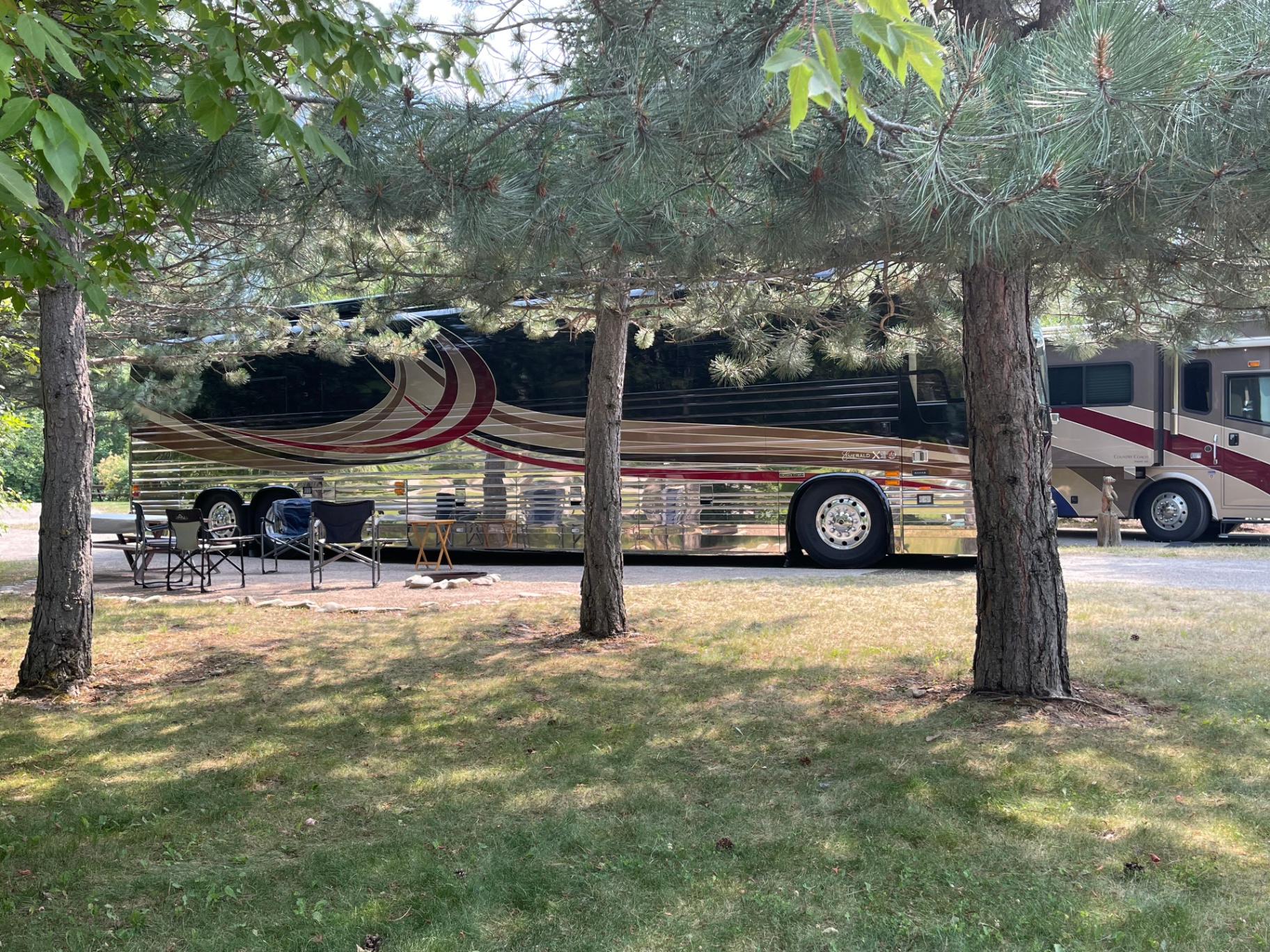 A luxury Prevost motorcoach with its awning extended and camp chairs set up on a shaded pull-through site at Campground St. Regis